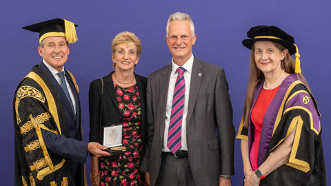 Four people stand against a purple background; two in academic regalia, one presenting a medal, and two in formal attire.
